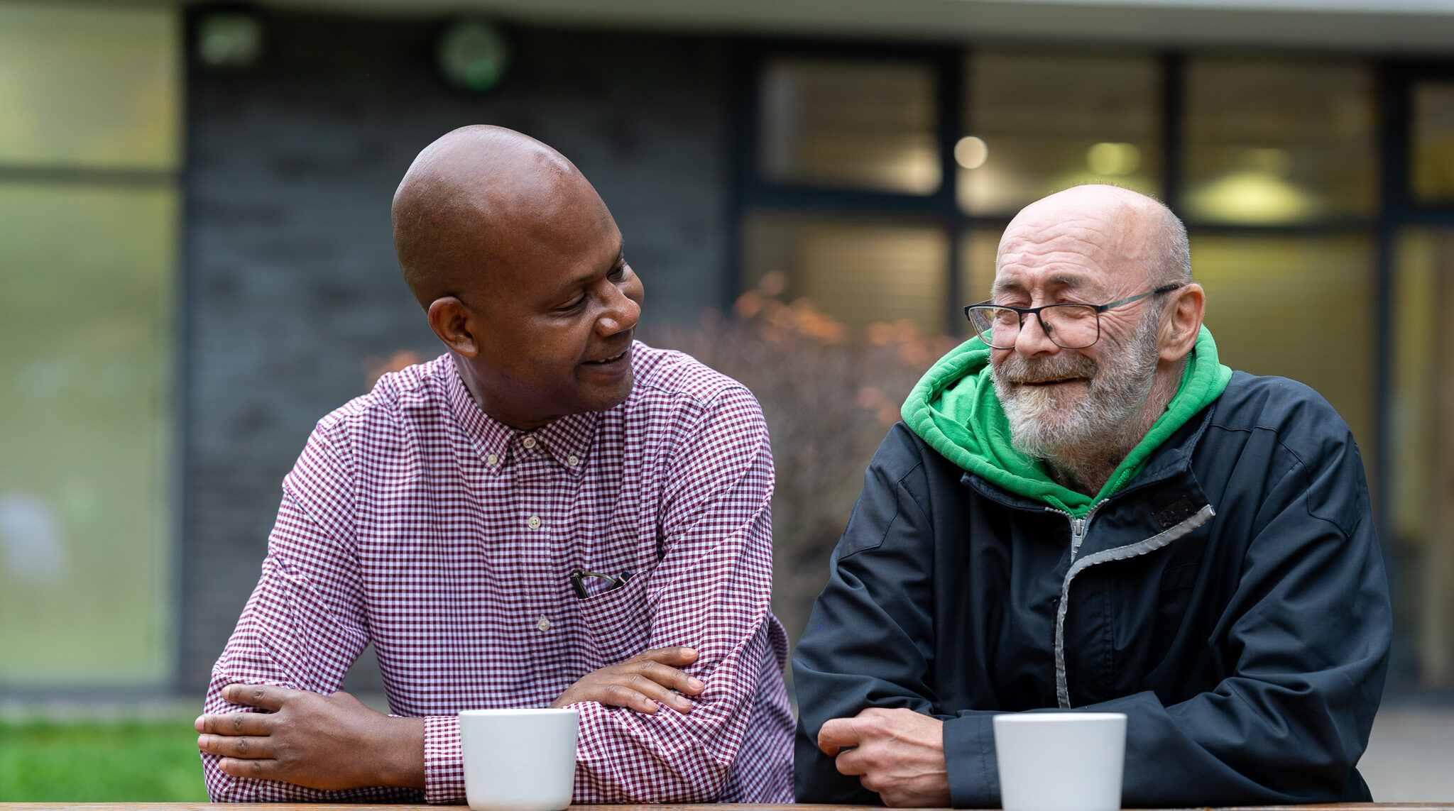 A support worker sits on a bench having tea with a customer at a Look Ahead homelessness and complex needs service.