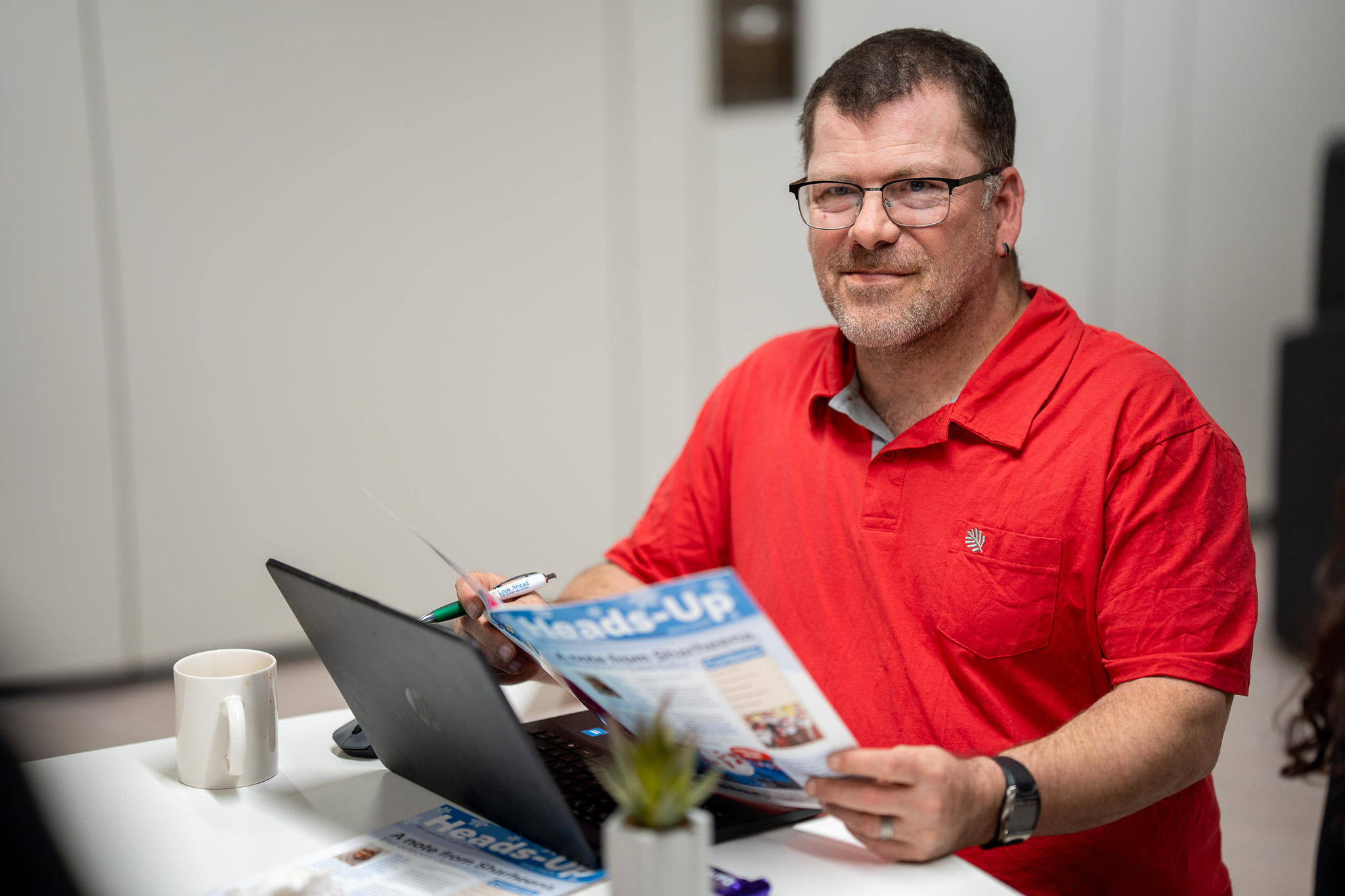 A man in a red polo shirt and glasses sat at a desk with a laptop and magazine.