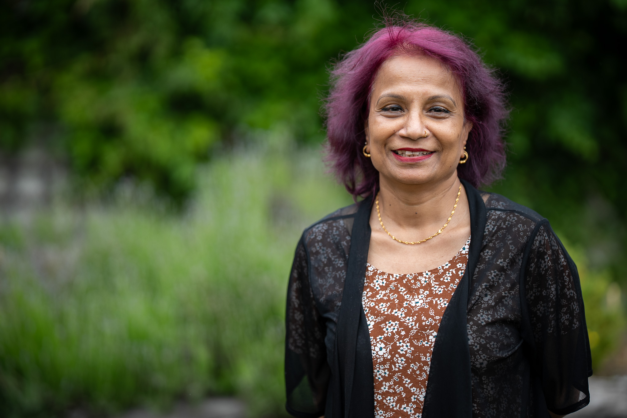 A smiling woman with purple hair, a reddish brown floral dress and black see through cardigan.