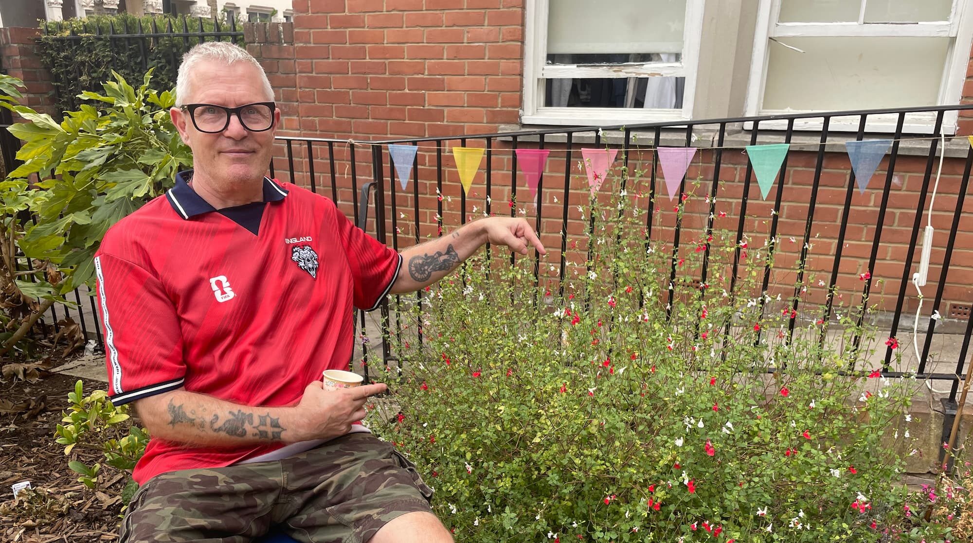 A man in a red England shirt and camouflage shorts with white hair sitting on a deck chair pointing towards a bush.