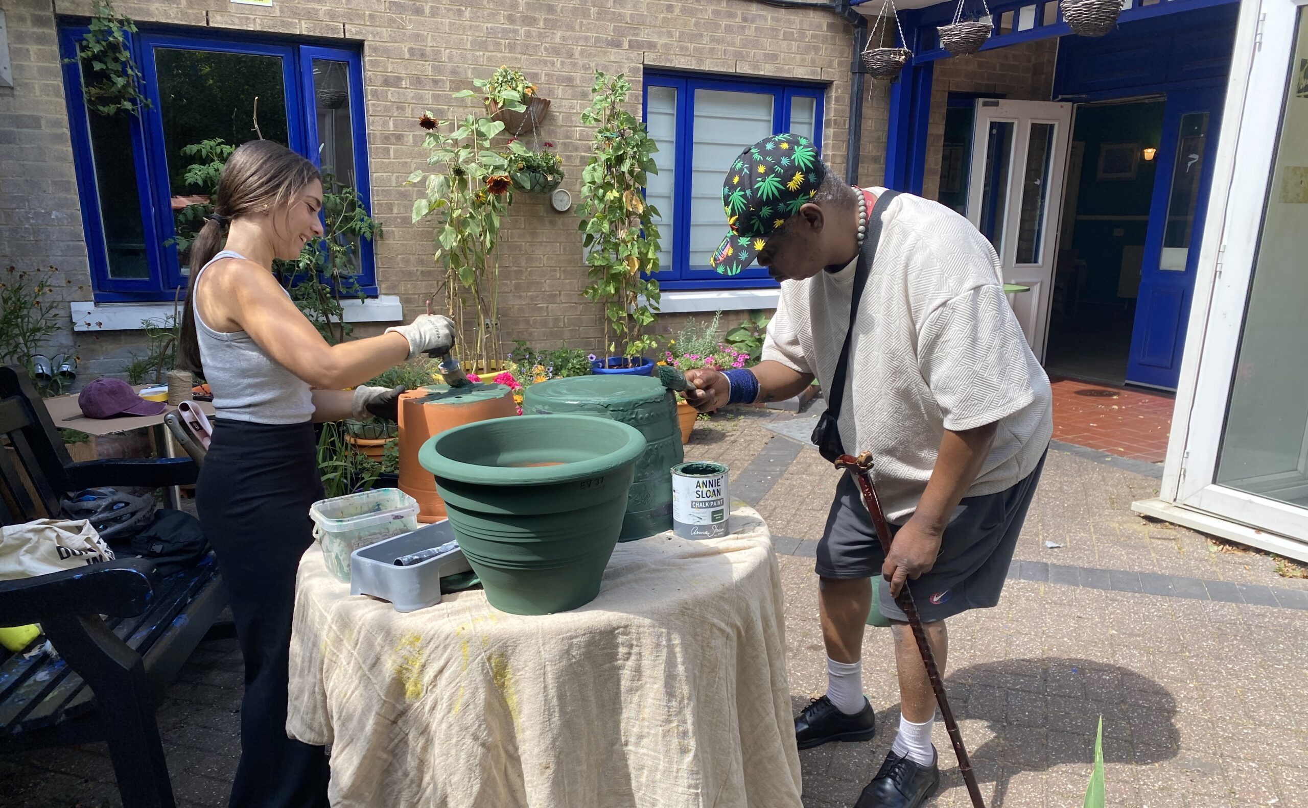 A lady in a grey top with her hair in a pony tail, and a man in a baseball cap with a walking stick, both are painting flower pots green.