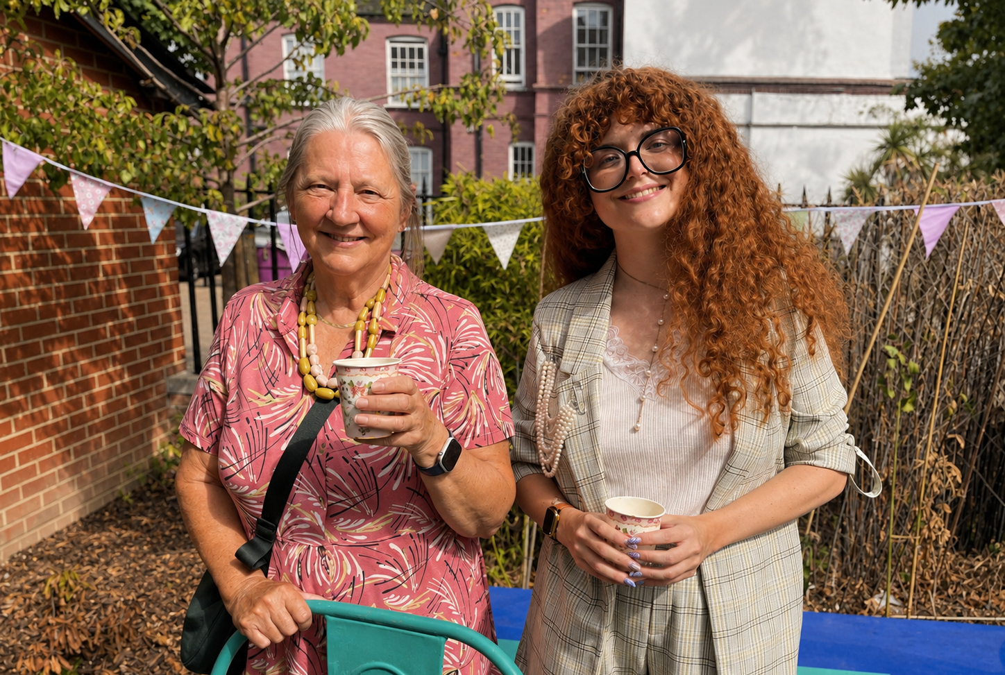 A lady in a pink floral dress holding a paper cup and another lady in a white t-shirt in a checked creamy green coloured jacket.