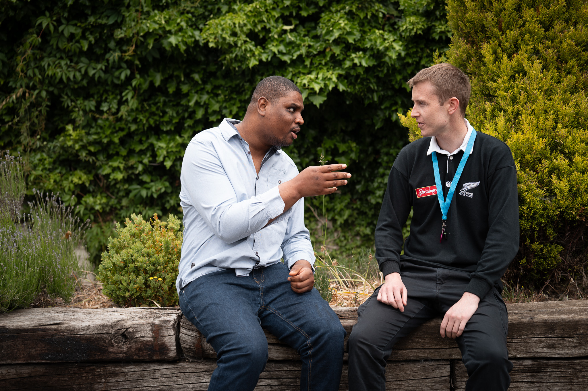 A man in a light blue shirt and jeans sits on a garden bench with a hedge in the background and chats to his support worker who is wearing a black New Zealand rugby shirt.