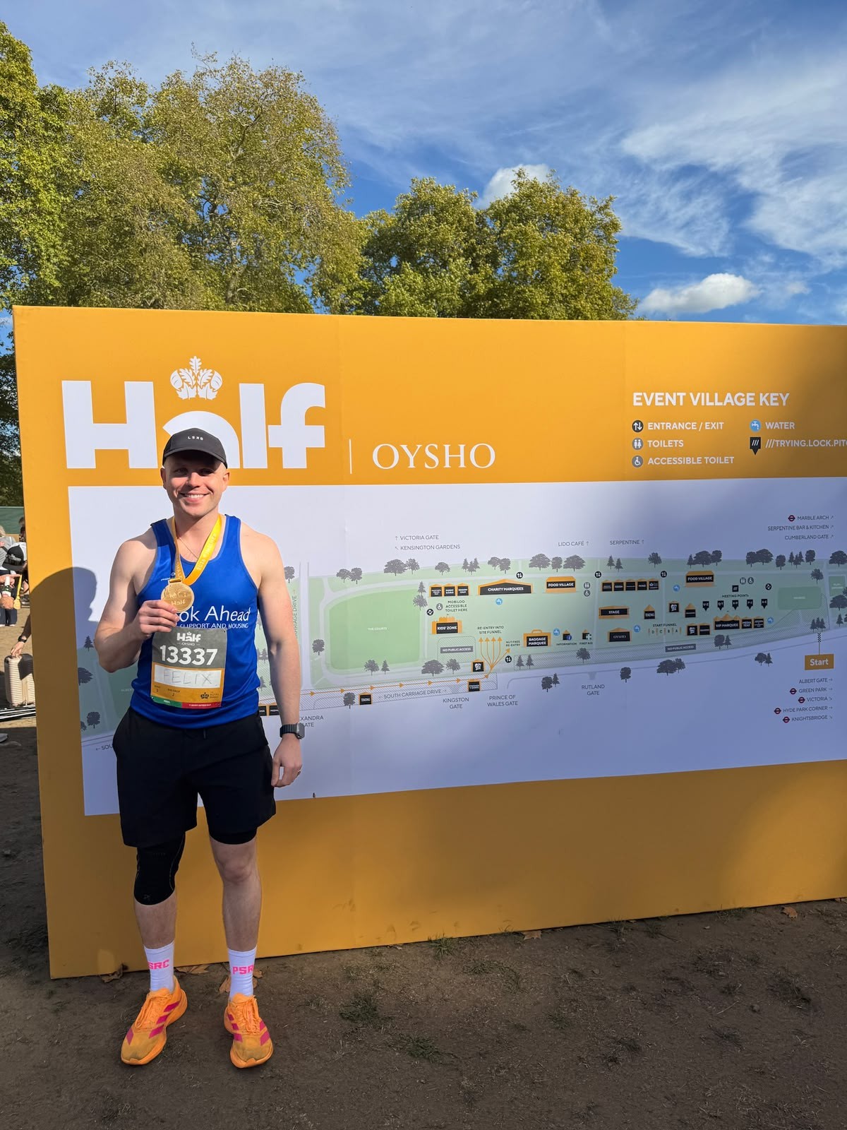 A man in a blue vest standing in front of a map of the Oysho Royal Parks Half Marathon Run, holding his medal.